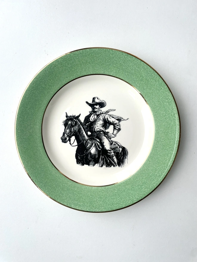vintage plate hanging on a white wall, it has a decorative border of grey and blue leaves, with a printed vintage illustration of a cowboy in the middle 