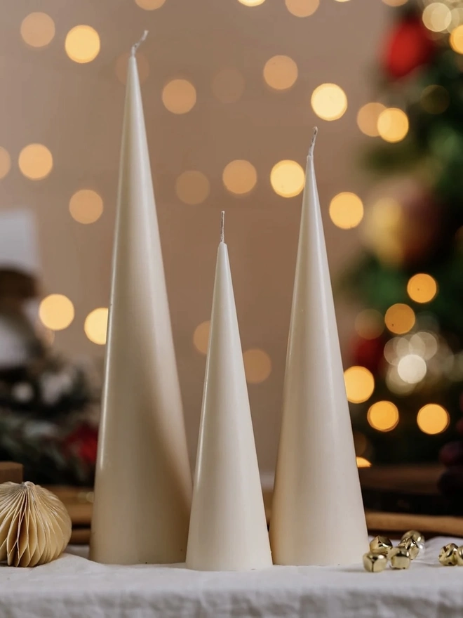 White Christmas Tree Cone Candles (Set Of 3)Three unlit cream cone candles in graduating heights, styled on white linen with a paper bauble and gold bells against festive bokeh lights. The sleek minimalist shape makes an elegant Christmas centrepiece.