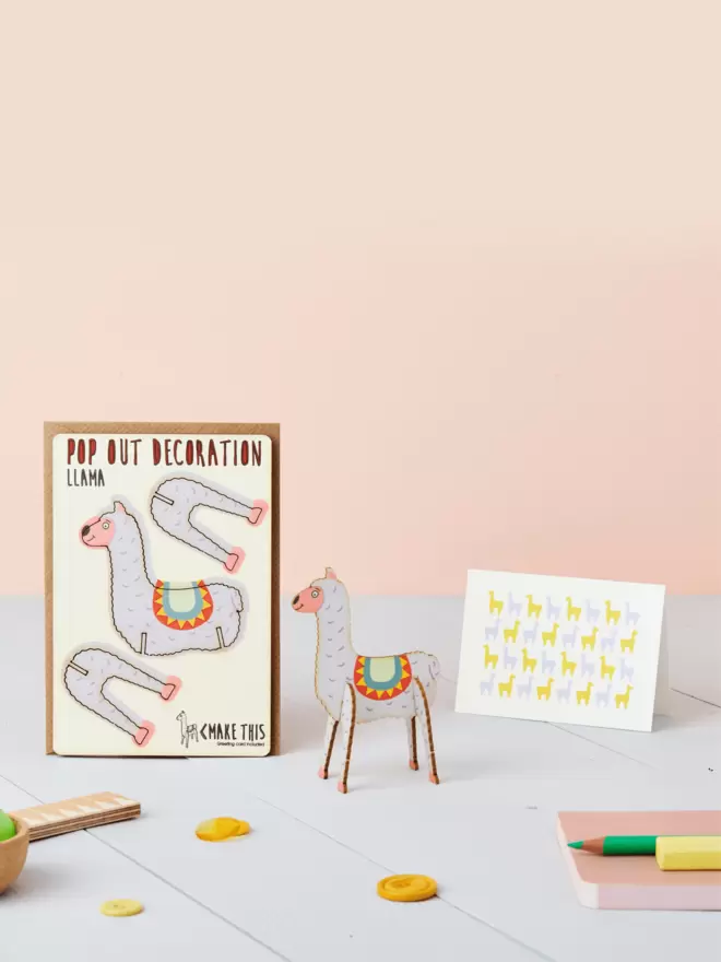 3D llama laser-cut decoration and llama pattern greeting card and brown kraft envelope on top of a wooden desk in front of a peachy pink coloured background