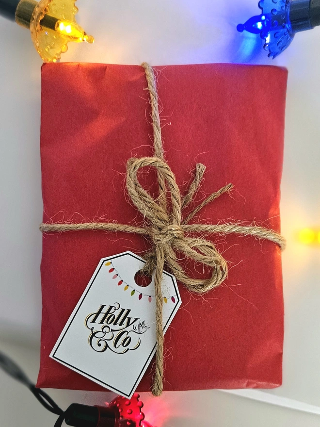 Photograph of the bauble wrapped in red tissue paper with brown twine and a gift tag illustrated with a colourful string of vintage baubles and the Holly and Co logo