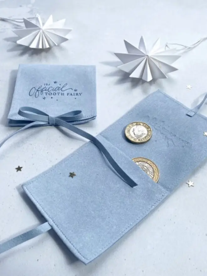 A pale blue pouch lies open with coins tucked into its inner pocket. A second pouch, tied with a bow, sits beside white paper stars.