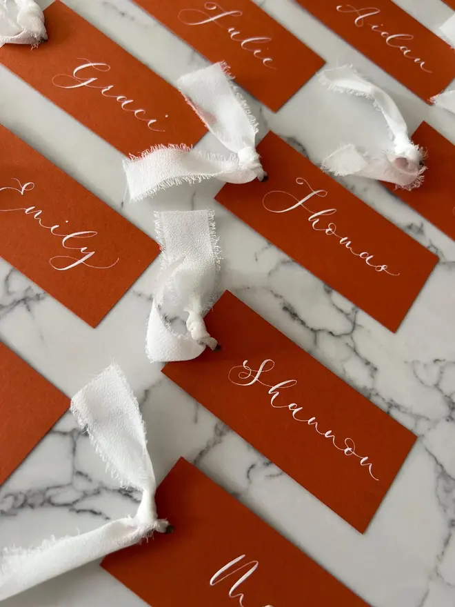 Personalised Calligraphy Rust Orange Place Card, orange place cards with white chiffon ribbons grouped together on a white surface. 