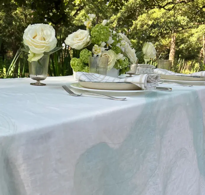 Imola Tablecloth. Light blue and white tablecloth with subtle abstract patterns, styled on an outdoor dining table with plates, glassware, and white floral arrangements.