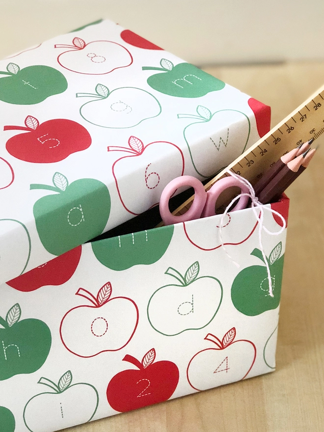 A gift wrapped in red and green apple wrapping paper, with letters and numbers inside each apple, rests on a wooden surface.