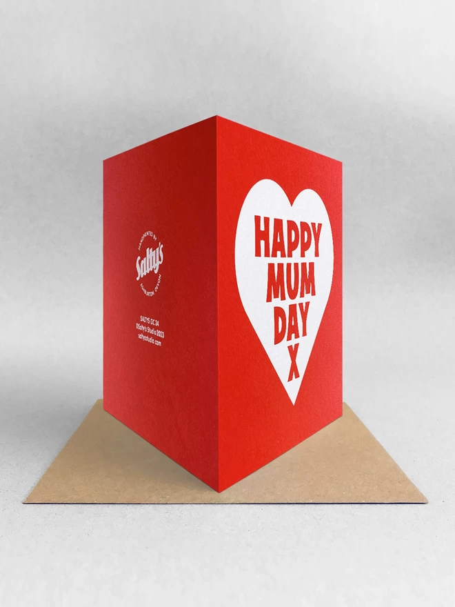 Rear view of a red square card with a white heart, containing the words Mappy Mum Day x. Stood on brown kraft envelope in a light grey studio 