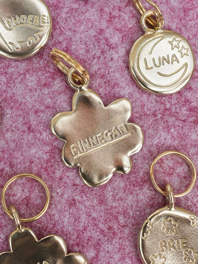 a bloom brass flower shape pet tag lays surrounded by bespoke custom name tags on a pink background