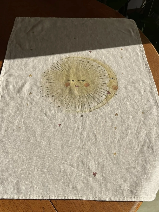 'Astral Weeks' Linen Tea Towel, a print sitting on a wooden surface with a sun motif in the centre. 