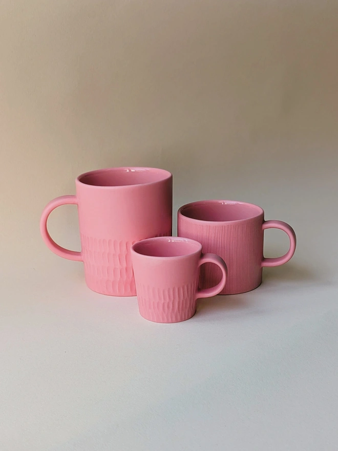 Three handmade porcelain mugs and coffee cups in Milkshake Pink. A large tea mug with a hand carved scalloped texture around the base, a small mug for flat white coffee which is hand carved in a combed texture, and espresso cup with scalloped hand carving around the base. All are glazed inside and matte outside.