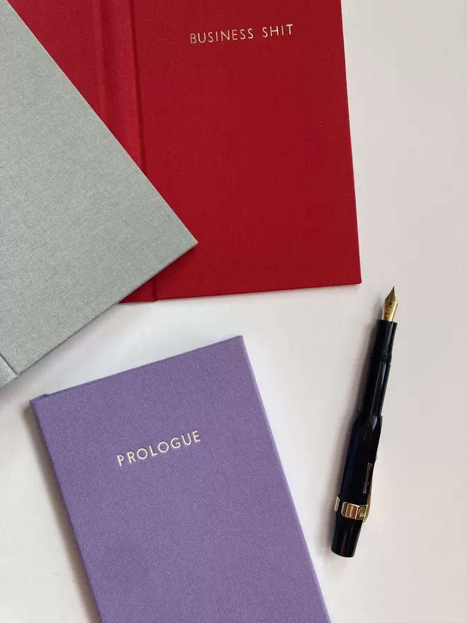 Three handmade pocket sized notebooks in peony red, crocus purple and dove grey book cloth with bespoke foiled lettering and fountain pen