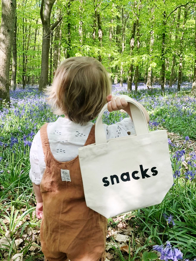a little boy holding up a mini kid's size tote bag with the word snacks on it