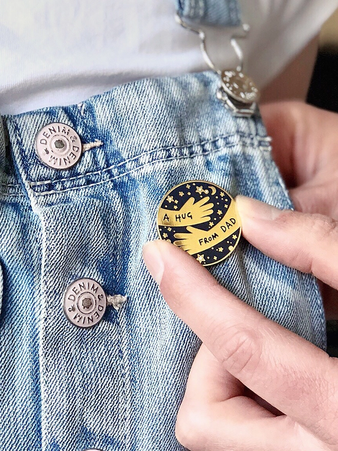A Dad is pinning a navy and gold hug from Dad pin badge onto child wearing denim dungarees 