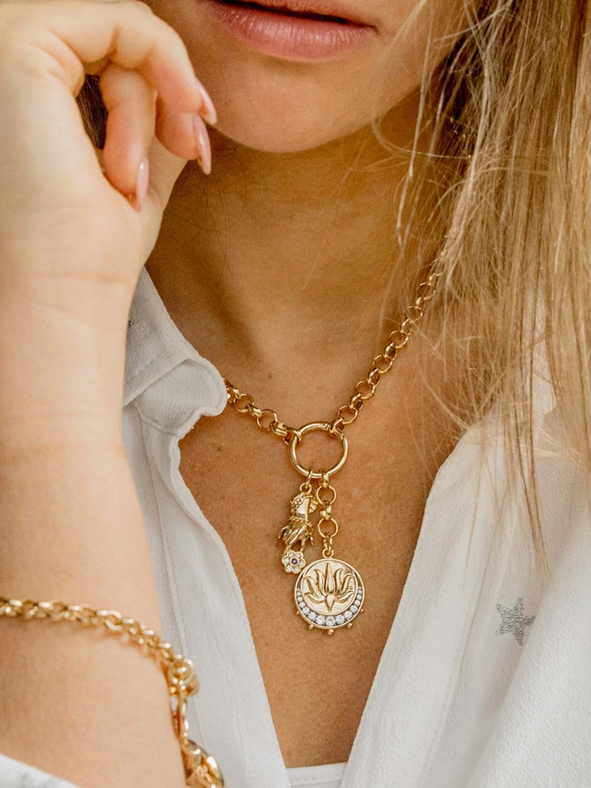 Woman wearing a white shirt with a gold belcher chain necklace hung with a gold lotus medallion charm and a gold hand charm