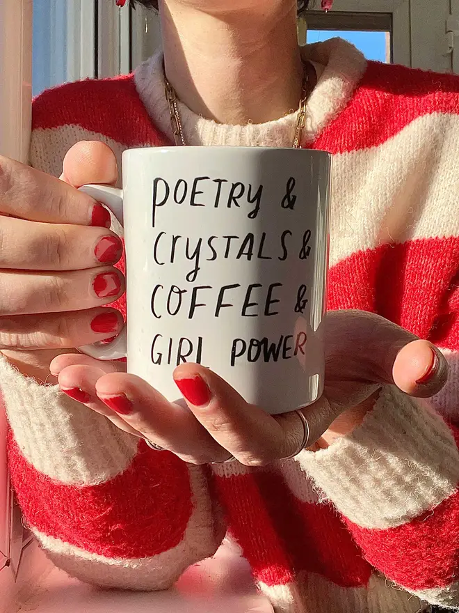 Nicola is holding mug which reads 'poetry & crystals & coffee & girl power' Sun shines through window and her nails are painted red