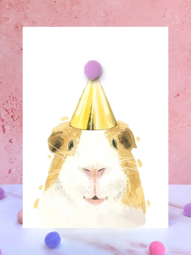 A greeting card featuring a hand painted design of a guinea pig, stood upright on a marble surface surrounded by pompoms. 
