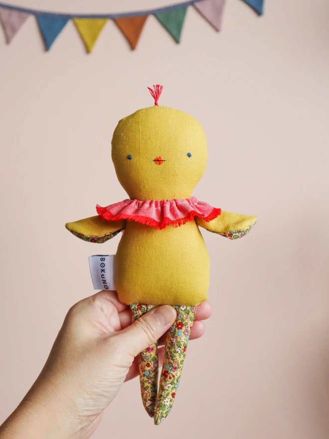 Handmade chick doll made from soft yellow fabric, with a stitched face, small beak, ruffled collar, and patterned fabric arms and long legs, held in a hand.