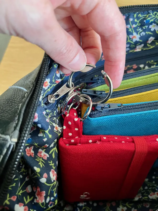 A hand opening a hinged silver ring attached to a set of colourful coin purses and a red card wallet.