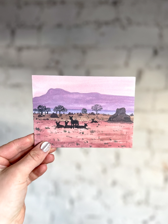 a greetings card featuring an illustration of a dusky coloured landscape and the silhouettes of some african wild dogs