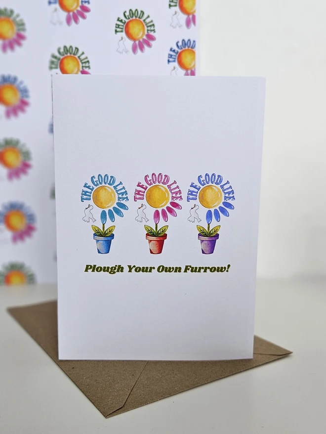 A white card with an illustration of  3 colourful flowers each in a terracotta pot displayed in a row with a white bird. The Good Life is written using a retro style font in 3 colours. It says at the bottom of the card Plough Your Own Furrow!
