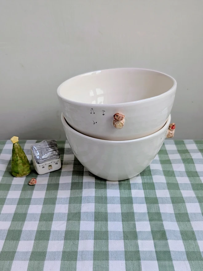 stack of two white ceramic bowls each with a tiny modelled robin perching on a modelled branch with musical notes resting on a green gingham tablecloth with a tiny pottery house beside them