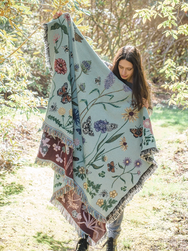 'Pollination Meadow' Recycled Cotton Blanket by Arcana seen held by a woman walking outside.