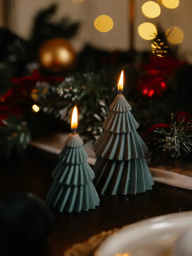 Forest Green Christmas Tree Warm Candles (Set Of 2). Two lit deep green Christmas tree candles with pleated, layered tiers glowing beside frosted pine garland and golden bokeh lights. The rich forest colour brings a classic festive feel to a mantelpiece or dinner table.