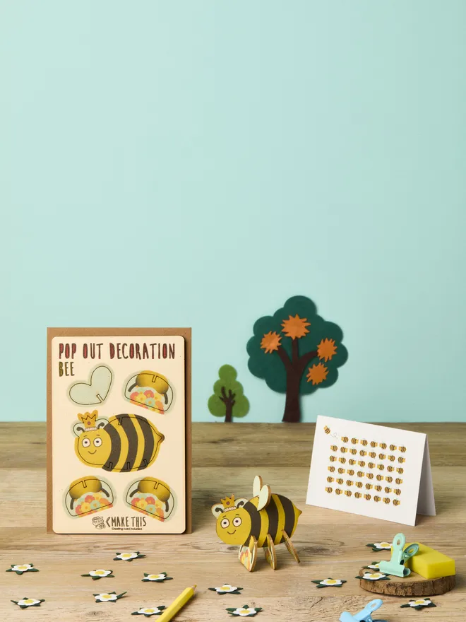 3D bee decoration and bee pattern greeting card and brown kraft envelope on top of a wood desk in front of a light blue coloured background