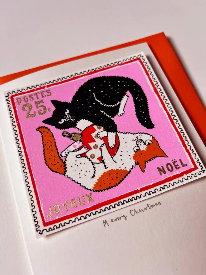 Happy Christmas Kittens Card, a white Christmas card with a colourful cat design in the centre. It is placed on an orange envelope on a white backdrop. 