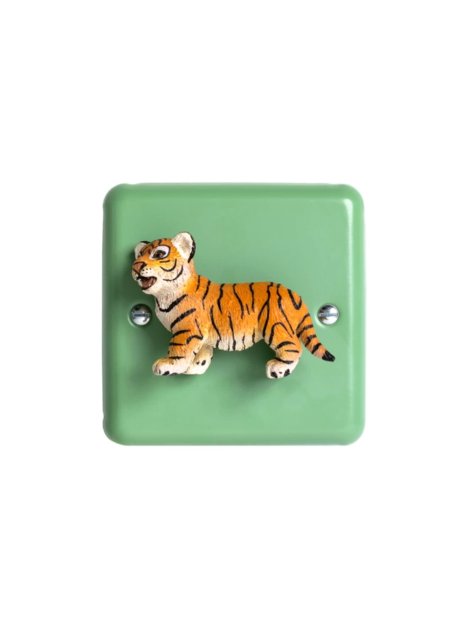 A tiger light switch. A pastel green nursery dimmer light switch with a tiger as the rotary knob to turn the lights on and off. The light switch plate is beryl green and made of metal, epoxy coated steel by Varilight. The tiger is made of plastic. The animal light switch brand is Candy Queen Designs.