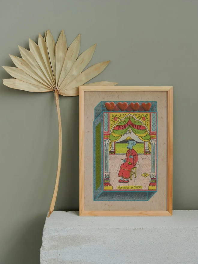 A surreal vintage-inspired matchbox illustration with the name “Fiona Apple” displayed on a red banner above a stage framed by ornate columns and green curtains. At center stage, a blue dog-headed figure dressed in a flowing red robe sits calmly on a wooden stool, holding a long smoking pipe. The tiled floor is patterned in pink, and the background opens to a stylized countryside landscape with striped green fields and a tiny thatched hut. Along the top edge of the box, five heart-shaped red match heads peek out, adding a playful touch. At the bottom, text reads: “Handmade by Caridade.” The overall effect is whimsical, theatrical, and dreamlike, blending animal fantasy with vintage ephemera.