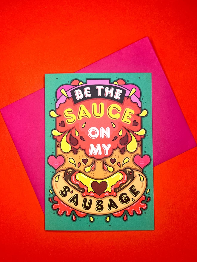 A green card featuring the phrase “Be the sauce on my sausage” on top of an abstract illustration including a hot dog, squirts of ketchup and mustard, and colourful hearts, sits on top of a pink envelope on a red backdrop.