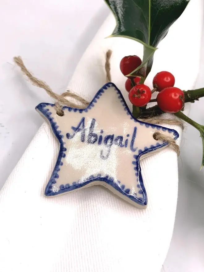 Personalised Ceramic Christmas Star Name Place Markers, a colourful ceramic start sitting on a white surface attached to a holly bush. 