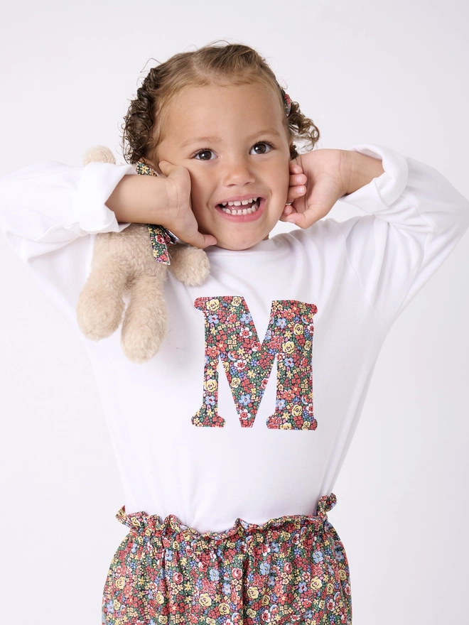 a 3 year old girl wearing a personalised kids white t-shirt with an initial cut from liberty floral print on the front