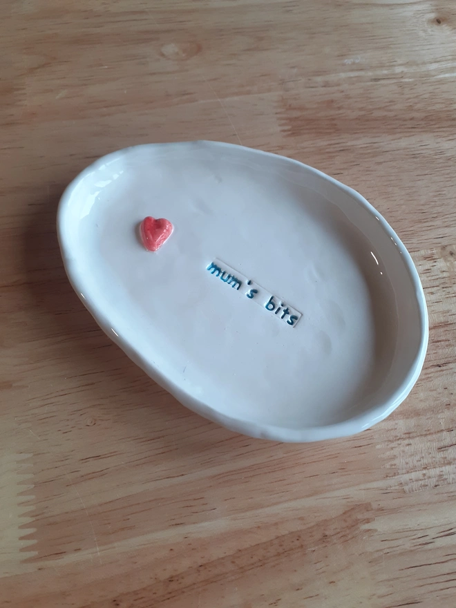 Handmade ceramic trinket dish on a wooden table, stamped with “mum’s bits” and finished with a small red heart, featuring slightly wonky oval shapes and glossy white glaze.