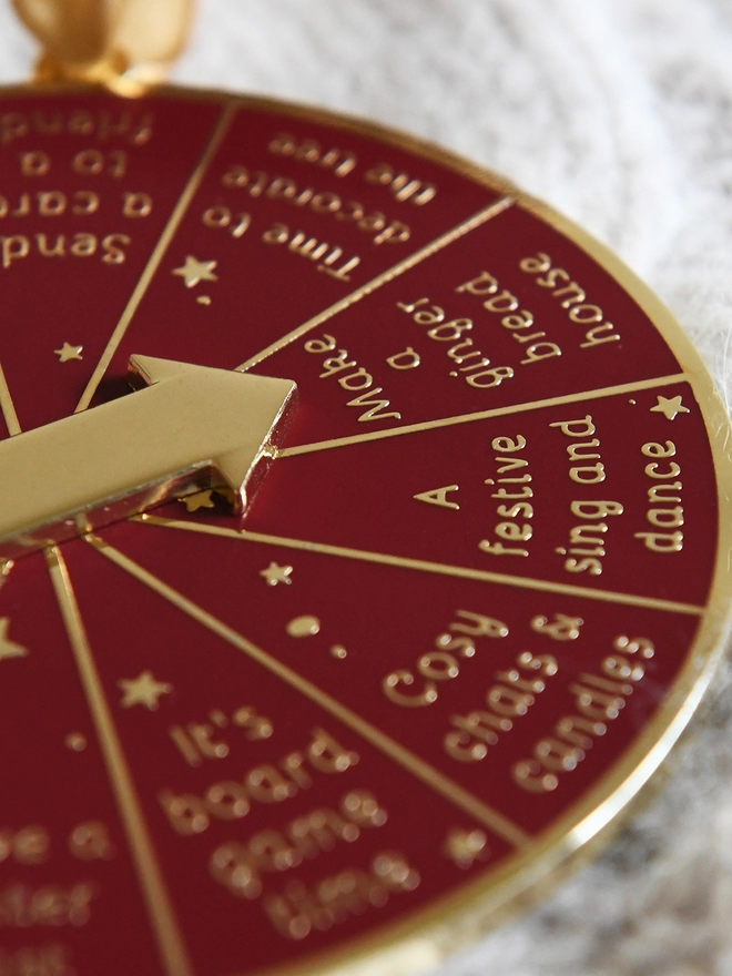 A deep red and gold Christmas decoration lays on a woollen blanket. It has 12 segments, each one with a different Christmas activity idea, and a golden arrow in the centre.