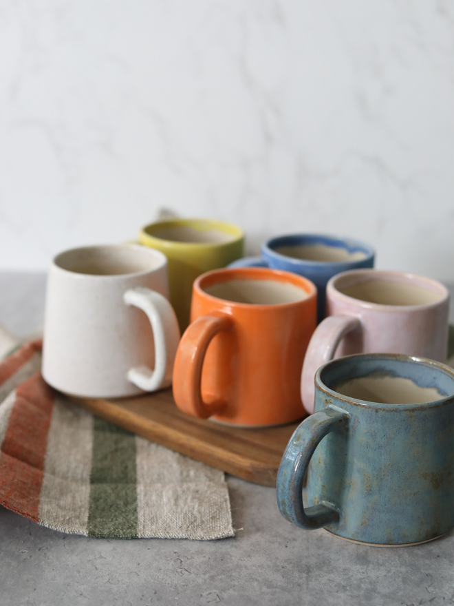 Six handmade ceramic standard mugs arranged on a wooden board viewed from a slight overhead angle, showing the full colour range of speckled white, yellow, mandarin orange, blue, blush pink, and teal, each containing a hot drink, styled with linen cloths against a white marble background