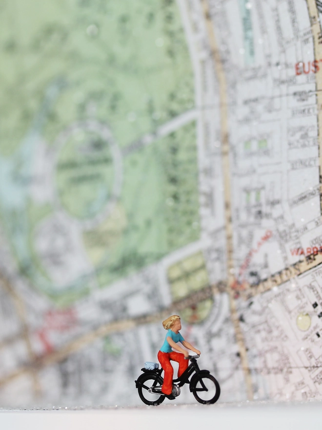 Artbox comprising a tiny woman on a bike cycling against sparkling map backdrop