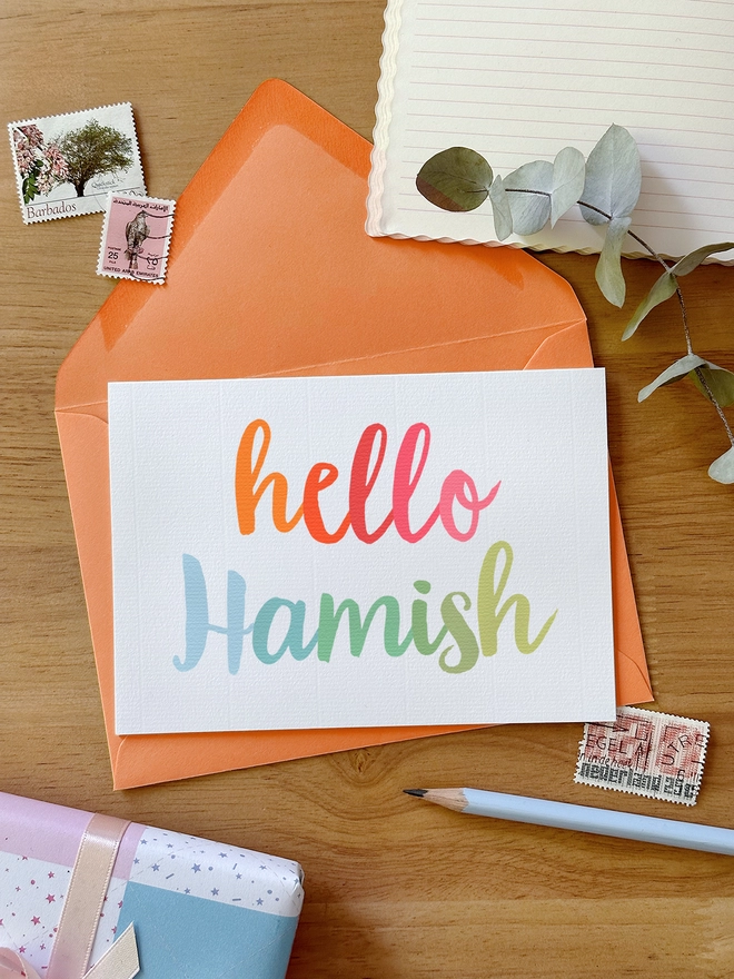 A personalised new baby greetings card with a name printed in pastel rainbow colours lays on an orange envelope on a wooden desk.