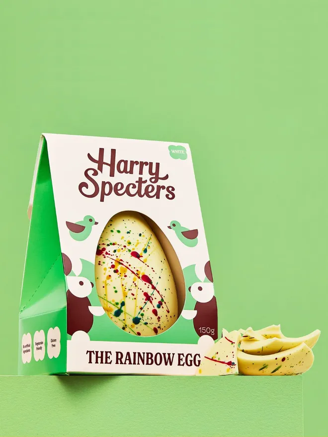 A white chocolate Easter egg decorated with rainbow speckles, next to a stack of white chocolate Easter egg pieces