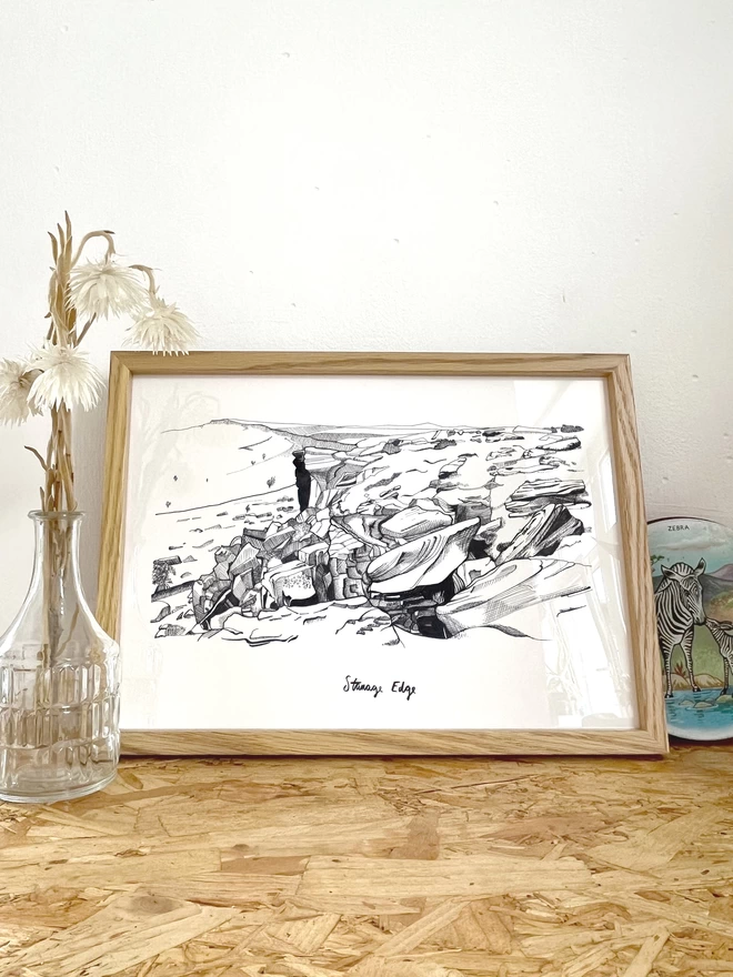a print featuring an fine line illustration of Stanage Edge in the peak district in a frame next to some flowers, a rock and a candle
