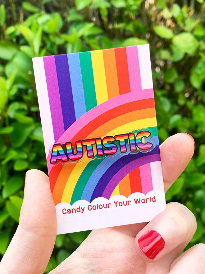 image shows a hand holding a rainbow business card. pinned to the card is an acrylic badge in the shape of the word 'autistic'. the letters are filled with horizontal rainbow stripes.