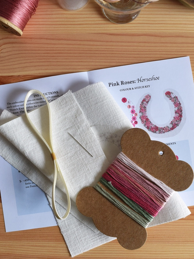 Embroidery Kit contents displayed on top of a pine chest.  Showing: thread card with coloured yarns, pre-printed design on linen, a length of ribbon, a needle, an extra linen and full instructions.