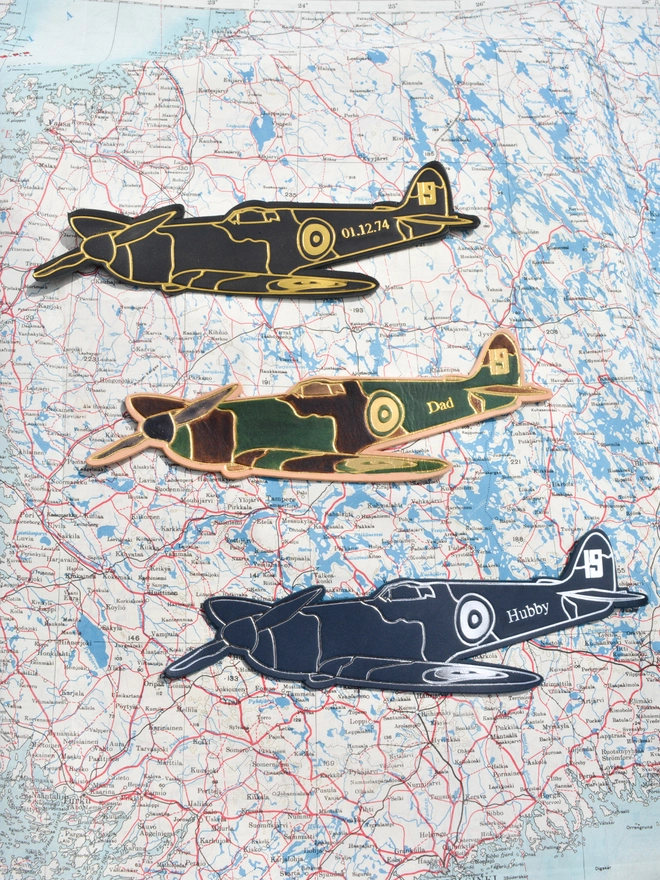 3 bookmarks in the shape of a World War Two spitfire airplane made in vegetable tanned leather in black and gold, blue and silver and a hand painted version in green and brown with personalisation
