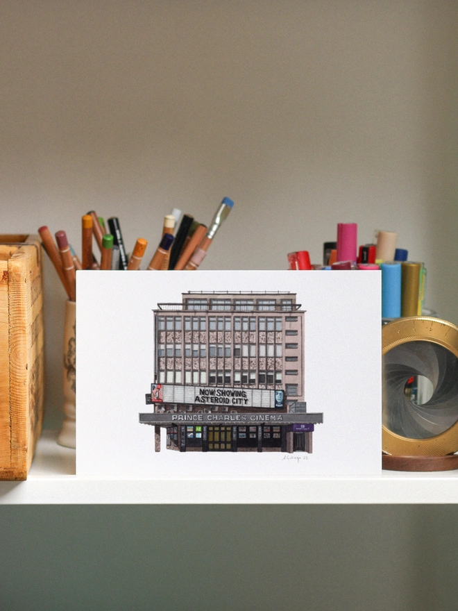 A watercolor print of the Prince Charles Cinema displayed on a shelf among various art supplies.