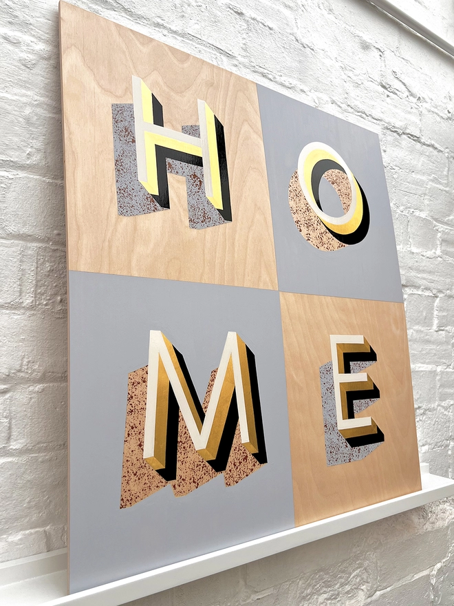 HOME hand painted sign in 23 carat gold leaf, pale blue, off white, tan, against a white brick wall, at an angle. 