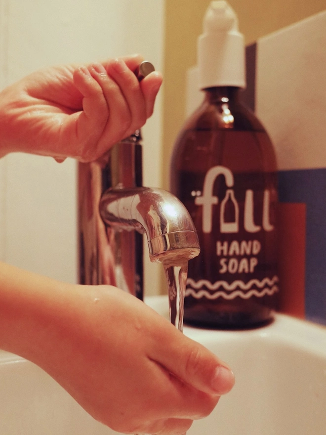 hands washing under silver tap with amber handsoap in background