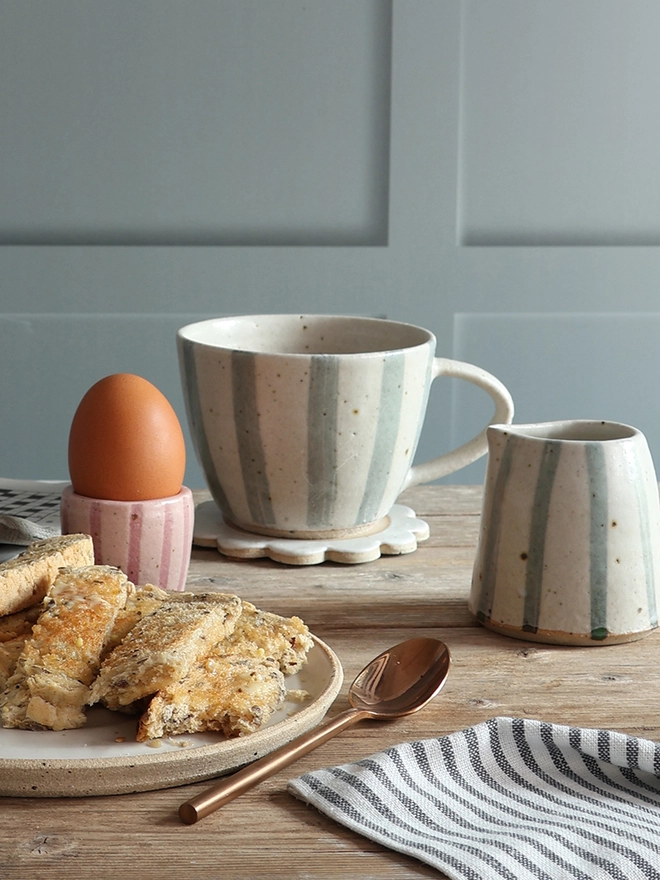 A table scene showing a pink stripe handmade ceramic egg cup holding a boiled egg, a mug and milk jug in blue and white stripe, and a plate with toast. A brass spoon and blue and white striped napkin can be seen in the foreground. 