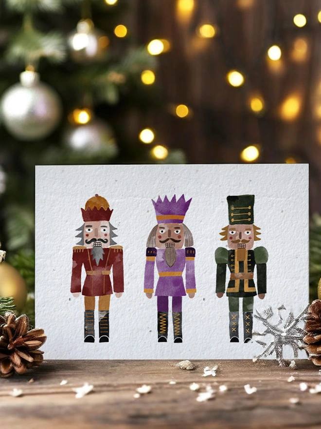 A landscape card featuring three watercolour nutcrackers, behind the card is twinkly lights and a Christmas tree
