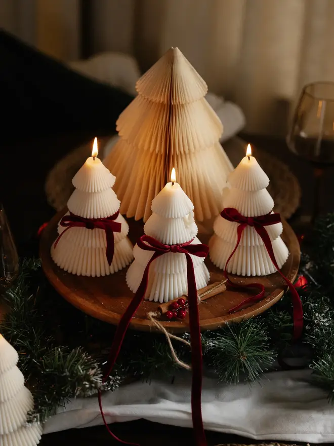 Tiered Christmas Tree Candle. Three lit cream Christmas tree candles with ribbed tiers and burgundy velvet bows, arranged on a wooden board in front of a paper honeycomb tree. Pine garland, cinnamon sticks and red berries complete this cosy festive centrepiece.