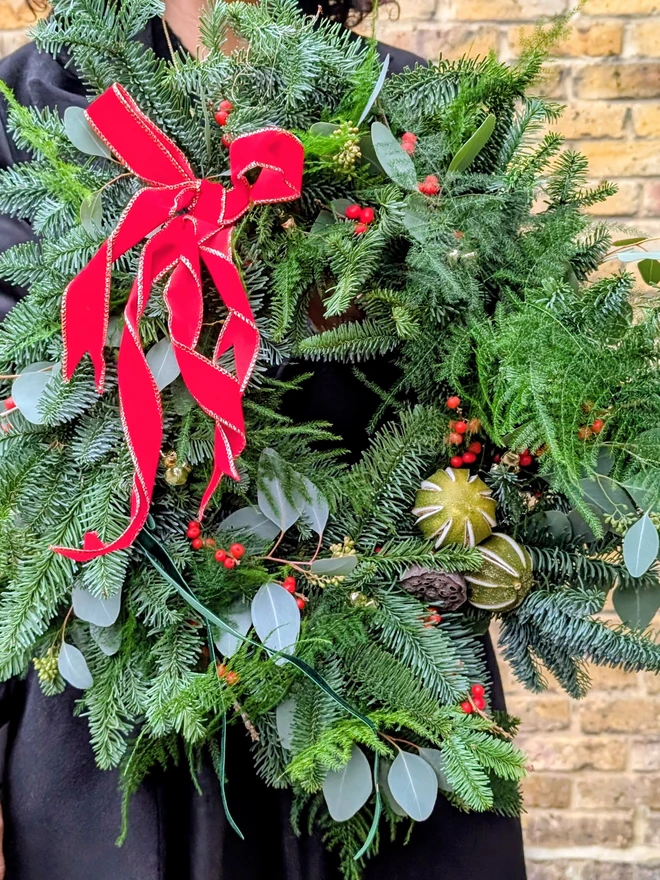 girl holding, Fresh handmade Christmas wreath with fir, eucalyptus, red berries, dried citrus, and natural seed pods, finished with a red velvet ribbon edged in gold  traditional festive front door decoration
