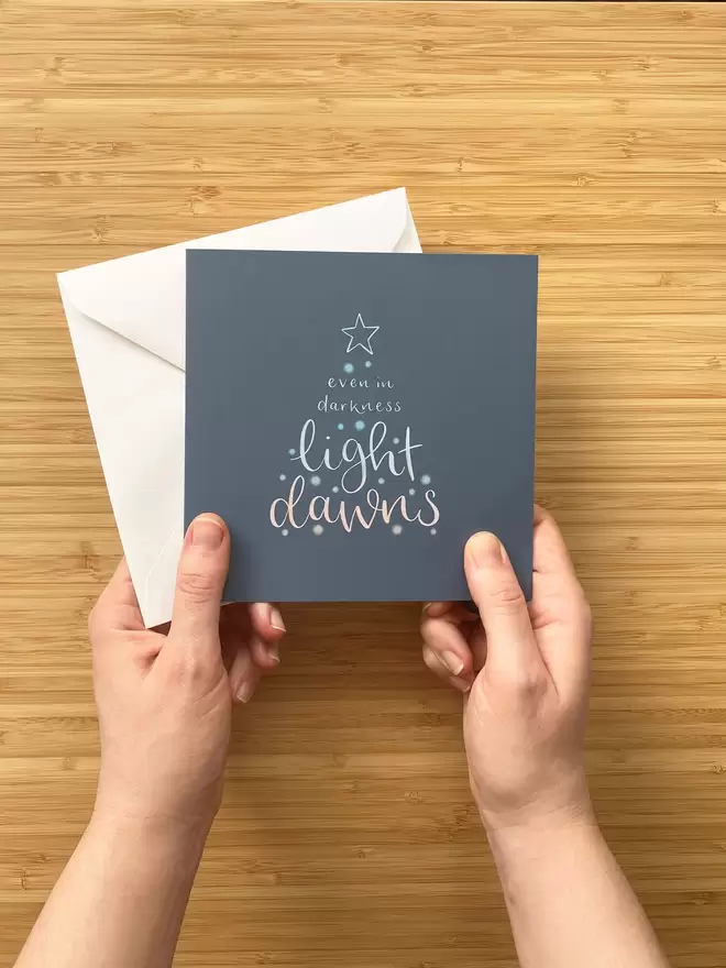 Square 'Light Dawns' Christmas card and white envelope, held in two hands by the designer